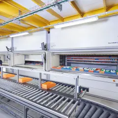 Ein Kommissionierlager mit automatisierten Lagersystemen. Eine Mitarbeiterin entnimmt Teile, während Behälter auf einem Rollenband transportiert werden.