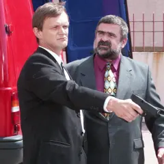 Gangster z filmu Kiler, w garniturze, celuje z pistoletu. Obok stoi mężczyzna w czarnym garniturze.
