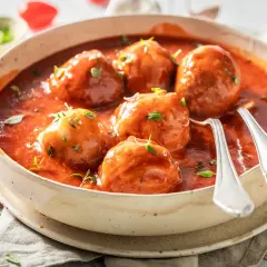 Albóndigas caseras con tomate frito, servidas en un cuenco de cerámica con hierbas frescas y dos cucharas listas para disfrutar.