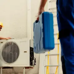 Técnicos revisan unidad exterior de aire acondicionado que dejó de funcionar de repente. Uno con casco amarillo examina la máquina, otro lleva una caja de herramientas azul.