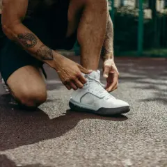 Mężczyzna z tatuażami na ramieniu klęczy, sznurując białe buty sportowe. Może to być przygotowanie do treningu lub ulga po bólu kolan po ćwiczeniach.