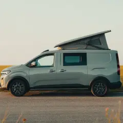 Un Dacia Sandman gris, prêt pour l'aventure, avec son toit relevable ouvert, stationné au bord d'un champ doré au coucher du soleil.