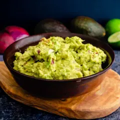 Miseczka guacamole z kawałkami cebuli i pomidorów, na drewnianej desce, w tle awokado i limonki.