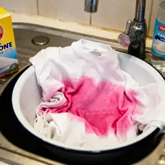 Un t-shirt blanc taché de rose trempe dans une bassine. À côté, une boîte de bicarbonate de soude et une bouteille de vinaigre, une astuce grand-mère pour le linge déteint.
