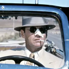 Jack Nicholson w kapeluszu i okularach przeciwsłonecznych, z plastrem na nosie, pali papierosa za kierownicą kabrioletu. Scena z filmu Polańskiego z Nicholsonem.