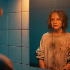 Mroczna scena z filmu "Morderczynie" - młoda kobieta w poplamionej bluzie patrzy w lustro, jej twarz wyraża niepokój.