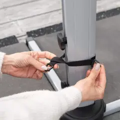 Dłonie zapinają czarny pasek na szarej podstawie parasola ogrodowego.