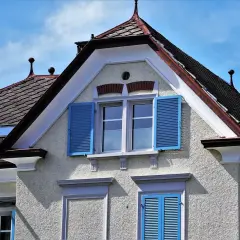 Elewacja domu z jasnym tynkiem, niebieskimi okiennicami i dachówką. Idealny grunt pod tynk zewnętrzny powinien być trwały i odporny na warunki atmosferyczne.