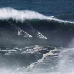 Surferzy ścigają się po gigantycznych nazare falach, tworząc białe smugi na ciemnym oceanie.