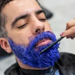 Un homme se fait teindre la barbe en bleu vif par un professionnel. La couleur est appliquée avec une brosse.