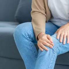 Frau sitzt auf Sofa, hält ihr Knie. Sie spürt ein unangenehmes Kribbeln in den Beinen.