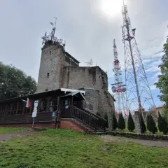 Kamienna wieża i drewniana przybudówka na szczycie góry. W tle wysokie maszty telekomunikacyjne. To popularne miejsce na szlaki turystyczne, jak Chełmiec.
