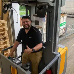 Mann gibt Daumen hoch in einem Hochhubwagen, bereit für die Stapler Unterweisung. Paletten stapeln sich links.