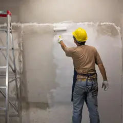 Operaio con casco giallo stende le vernici bianche su un muro grezzo, accanto a una scala metallica.