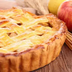 Tarta de manzana dorada con rejilla de masa, junto a manzanas frescas y palitos de canela.