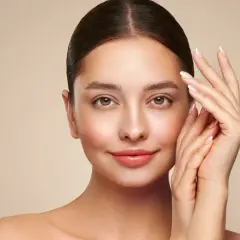 Portret kobiety z gładką cerą i dłońmi przy twarzy na beżowym tle.