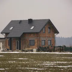 Dom w budowie z cegły, z szarym dachem i oknami. W tle las. Czekamy na uprawomocnienie pozwolenia na budowę.