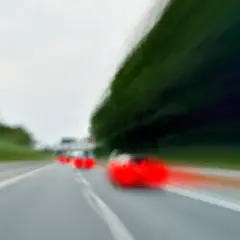 Rozmyte czerwone samochody na drodze, jeden z nich wykonuje wyprzedzanie z prawej strony.