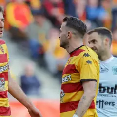 Piłkarze Jagiellonii Białystok w żółto-czerwonych strojach rozmawiają na boisku. W tle kibice, ważny mecz dla rankingów Jagiellonia Białystok – Piast Gliwice.