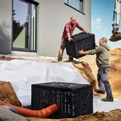 Zwei Männer installieren eine Sickerbox für die Sickerung des Hauses. Ein Bagger ist im Hintergrund zu sehen.