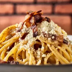 Pyszny makaron włoski carbonara, posypany parmezanem i chrupiącym boczkiem, na tle ceglanej ściany.