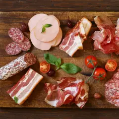 Assortiment de différents types de charcuterie : saucisson, jambon cru, bacon, mortadelle, accompagnés de tomates cerises et d'olives.