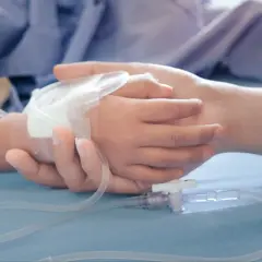Dłoń dorosłego obejmuje dłoń dziecka z wenflonem. W tle pluszowy miś. Wsparcie w trudnych chwilach, jak przy ubieganiu się o **zasiłek pielęgnacyjny**.