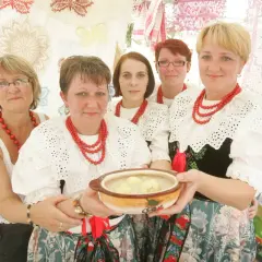 Pięć kobiet w tradycyjnych strojach ludowych prezentuje miskę z zupą. Jedna z nich trzyma ją w dłoniach. W tle widać ozdobne koronki, niczym z kukiełkowego teatru.