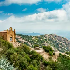 Pequeña ermita en la cima de una colina, con vistas a las impresionantes rutas de Montserrat y un paisaje montañoso bajo un cielo azul con nubes.