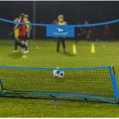 Piłkarski rebounder Yakimasport na zielonej murawie, z piłką w siatce. W tle trening młodych piłkarzy.
