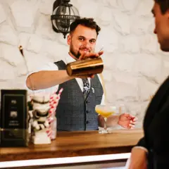 Uśmiechnięty barman w kamizelce nalewa drinka do kieliszka.