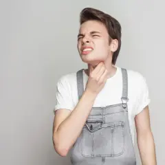 Młody mężczyzna w ogrodniczkach trzyma się za gardło, czując nieprzyjemne uczucie guli w gardle.