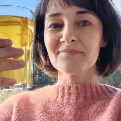 Kobieta trzyma szklankę z napojem, być może to picie pokrzywy, ciesząc się widokiem na las. Efekty zdrowotne napoju są widoczne w jej promiennej cerze.