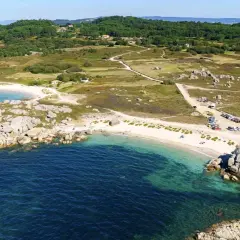 Vista aérea de una playa con arena blanca y aguas turquesas, rodeada de rocas y vegetación. Se ve un aparcamiento con coches cerca de la playa.