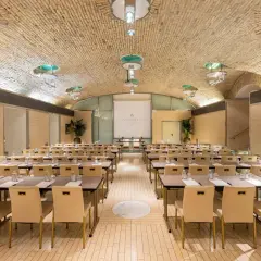 Sala riunioni con tavoli e sedie disposti per un evento all'Anantara Palazzo Naiadi Rome Hotel. Soffitto a volta in mattoni e illuminazione moderna.