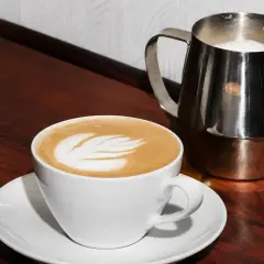 Biała filiżanka z latte art, obok metalowy dzbanek z mlekiem. Idealny raf coffee na drewnianym blacie.