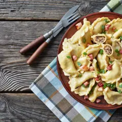 Pyszne pierogi z mięsem, posypane skwarkami i szczypiorkiem, czekają na talerzu. Obok sztućce i miseczka śmietany.