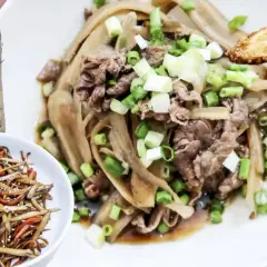 A delicious dish of braised burdock with thinly sliced beef and green onions, served on a white plate. A small bowl of burdock stir-fry is on the side.