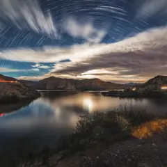 A surreal moon landscape with star trails arching over a calm lake and rugged mountains. Light trails from cars streak across the scene.