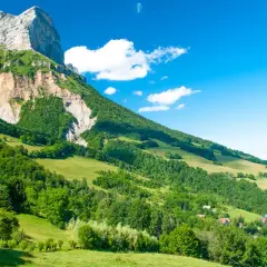 Vue panoramique d'une montagne imposante, le GR 965, dominant une vallée verdoyante parsemée de maisons. Le ciel bleu est parsemé de quelques nuages blancs.