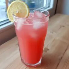 Ett glas rosa rabarberlemonad med is och en citronskiva, redo att avnjutas vid fönstret.