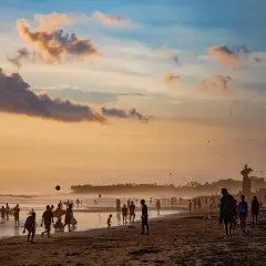 Zachód słońca na plaży Bali. Ludzie spacerują, grają w piłkę i puszczają latawce. W tle widać palmowy las.