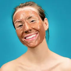 Ragazza sorridente con una maschera all'argilla sul viso, che promette un viso lucido e radioso.