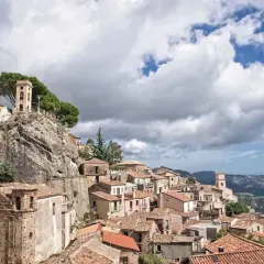 Charming towns in Calabria, Italy, cling to a rocky hillside under a dramatic sky. A bell tower stands sentinel over terracotta roofs.