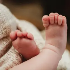 Delikatne, różowe stópki niemowlaka z lekko podwiniętymi palcami u nóg, otulone miękkim, beżowym kocykiem.