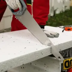 Ręce w rękawiczkach tną styropian piłą ręczną. W tle widać miarkę i materiały budowlane.