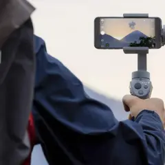 Osoba trzyma smartfon na gimbala, nagrywając piękny krajobraz z górami i zachodzącym słońcem.