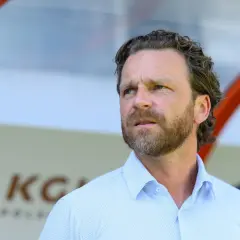 Bartosz Grzelak trener zamyślony, w jasnej koszuli, na tle stadionu.