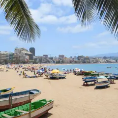 Solig strand i Las Palmas med palmer, båtar på sanden och en livlig stad i bakgrunden.
