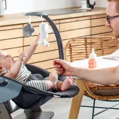 Ein Vater spielt mit seinem Baby in einem Hochstuhl. Ab wann Babys im Hochstuhl sitzen, hängt von ihrer Entwicklung ab.